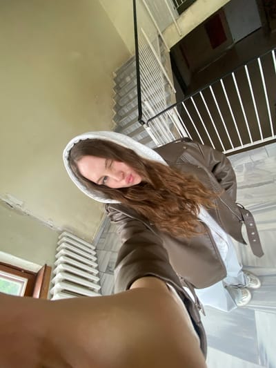 The image displays an indoor scene in the city of Марковский, Russia, captured from a high-angle selfie perspective. A young woman with long, wavy reddish-brown hair is visible, winking with her left eye and puckering her lips. She wears a white hooded sweatshirt beneath a brown faux leather jacket, light-colored trousers, and white sneakers. The background reveals a multi-level interior, likely a stairwell or hallway. Walls are uniformly pale beige with visible surface wear and minor discoloration. A white, multi-finned radiator is positioned below a brown-framed window, showing a glimpse of green foliage outside. The floor is tiled with a light grey and white marbled or patterned surface. A black railing with vertical white balusters separates an upper level or landing, adjacent to a staircase featuring light-colored steps.