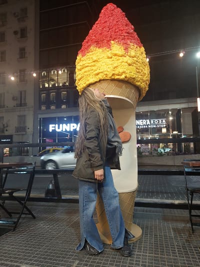 This vertical image captures an urban street scene at night in Buenos Aires, Argentina. The main subject is a person, appearing to be a young woman, standing next to a very large, oversized ice cream cone sculpture.
The person has long, light brown hair with highlights, wearing a dark olive-green jacket, blue jeans, and dark athletic shoes. A grey scarf is draped around her neck. She is looking upwards, towards the "scoops" of the ice cream cone.
The prominent ice cream sculpture features a textured red top scoop, blending into a yellow bottom scoop. The cone itself is a light brown, patterned with a waffle design. It appears to be an outdoor decoration or advertisement.
In the background, illuminated buildings line the street. One building clearly displays the text "NERA ROSA CASERA NON-STOP," suggesting a business specializing in homemade goods. Another building features a large, glowing sign that reads "FUNPA." Below, a smaller sign indicates "ilovegifts.com.ar," which confirms the Argentine location.
The street is dark, with artificial lights from the buildings and streetlights illuminating the scene. A blurred white car and a yellow vehicle (likely a taxi) are visible in motion on the street, indicating active city life. The ground is paved with dark, square-patterned tiles. Dark metal structures, possibly railings or outdoor seating, are visible to the right. The person's jacket and scarf suggest the weather might be cool.