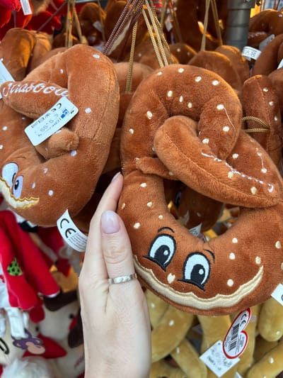 A close-up shot shows a person's hand holding a plush pretzel toy with embroidered white dots and a smiling face. The pretzel is a warm brown color and appears to be a Christmas ornament. It is hanging amongst other similar plush pretzels. A price tag is visible on one of the pretzels, reading "6.99" and "CE". In the background, other plush toys, including red and white characters, are visible. The scene is likely a Christmas market in Strasbourg, France, given the characteristic style of the plush toys and the presence of the "CE" marking.