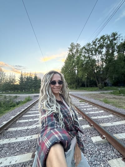 A young woman is captured in a selfie or portrait outdoors, standing beside train tracks during either sunrise or sunset. The image is initially oriented vertically but appears rotated 90 degrees counter-clockwise, showing the subject and landscape sideways.
The subject is a woman with long, wavy blonde or light brown hair, possibly featuring braided elements. She wears round, dark-lensed sunglasses and a plaid flannel shirt with patterns of red, dark blue, and white. She is smiling directly at the camera, conveying a cheerful expression. Her left arm is visible, resting by her side.
The setting is an open, natural environment dominated by train tracks. The tracks, composed of metal rails, concrete sleepers (ties), and gravel ballast, stretch into the background. To the left of the tracks, a strip of grass and sparse vegetation gives way to a dense line of trees, suggesting a wooded area or forest. A distant house or building is faintly visible through the trees. Overhead, several thin utility lines or power lines cross the sky.
The sky, a mix of clear blue and scattered clouds, is illuminated by soft, warm orange and yellow tones, characteristic of either dawn or dusk. The overall lighting is gentle, contributing to a serene atmosphere. On some of the concrete train ties, faint textual or numerical markings, such as "1994" and what appear to be Cyrillic characters, are visible, likely manufacturing stamps. The scene depicts a rural or semi-rural location in Rusanovo, Russia.