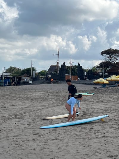 In North Kuta, Indonesia, on a cloudy day at the beach, two individuals are engaged in surfing activities. A person in a black long-sleeved wetsuit stands behind a person wearing a light blue long-sleeved rashguard and black bottoms, who is positioned on a blue surfboard. The person on the surfboard is bending over, holding onto the board, and appears to be in the process of standing up. Another surfboard, light beige in color, lies on the sand in front of the blue surfboard. The beach is sandy, with tire tracks visible in the sand. In the background, there are several structures including a low-rise building with a thatched roof, a wooden shack, and a stone staircase leading up to a building with a dark, textured facade. Yellow umbrellas and lounge chairs are set up further in the background, suggesting a beachside establishment. The sky is filled with grey and white clouds.