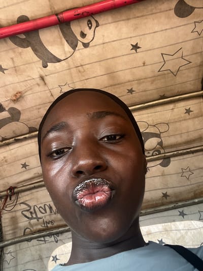 The image is a close-up, low-angle selfie of a young Black woman in Galadimawa Village, Nigeria. She is wearing a black hijab and a light blue shirt. Her lips are pursed and appear to be covered in glitter or a shimmery lip gloss. She is wearing small stud earrings. The background is a fabric material with a pattern of pandas and stars, and some horizontal lines that suggest ruled paper. A red rod is visible at the top of the image, and metal bars run horizontally across the frame. Some wires are also visible on the left side. The lighting is bright and appears to be natural, possibly from sunlight.