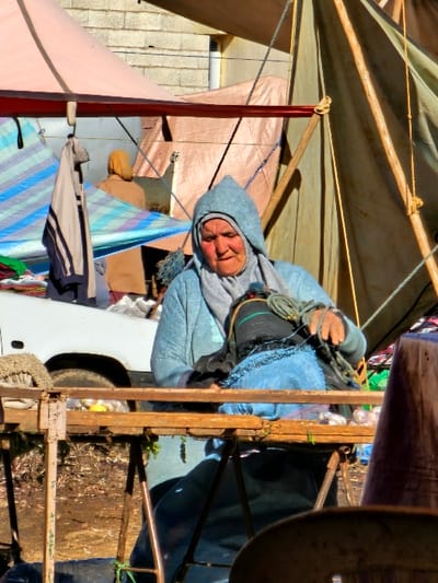 A person in a hooded blue jacket sits amidst a cluttered, makeshift structure. Ropes are bundled in their hands. Part of a vehicle is visible to the right. Assorted materials, including tarpaulins and cloth, form the roof and sides of the enclosure. The setting suggests a temporary or improvised living space.