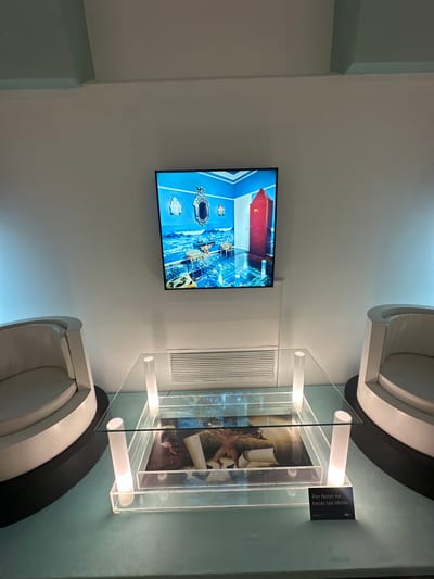Indoor scene showcasing an art display. A flat-screen TV mounted on a white wall displays an image of an interior with a red-ceilinged room and ornate furniture, potentially an artistic creation. Beside the TV is a ventilation unit or decorative panel. The display includes a clear, box-shaped showcase with white cylindrical supports, filled with printed materials or artwork. A small sign is attached to the showcase, bearing the Spanish text "Por favor no tocar las obras" (Please do not touch the works). Two white, semi-circular chairs with black bases flank the showcase. The color scheme is predominantly white, light blue, and gray. The location seems to be a museum or art gallery in Buenos Aires, Argentina. The display is focused on art and interactive viewing via the television screen. No people are directly visible in the frame.
