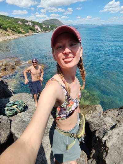 A woman in a floral bikini top and green shorts is taking a selfie with a lake in the background. She is wearing a red baseball cap and has her tongue sticking out. Behind her, a man in sunglasses and blue swim trunks stands on the rocky shore, holding his shorts. The lake is clear and blue, with some houses and green hills visible on the distant shore. There is a pile of green gear on the rocks in the foreground. The location is Utios, Ukraine.