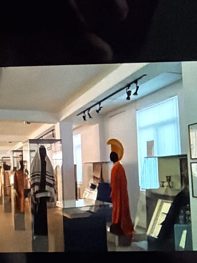 This media file depicts an indoor scene, possibly a retail environment or a museum, given the display of items on shelves and wall-mounted racks. The primary subject is a mannequin dressed in a vibrant orange garment, which appears to be a robe or a cape. The mannequin is positioned to the right of the frame, with its head tilted upwards and its body angled to face a lighter area of the room.
To the left of the orange-clad mannequin, a wall features a series of dark, stylized hooks, suggesting a place where garments or accessories might be displayed or hung. Further down this wall, a sequence of dark, abstract shapes resembling figures or sculptures are mounted, adding an artistic element to the setting.
The background reveals architectural features like a skylight or a large window, casting a bright, diffused light into the space, which is consistent with daytime. The scene is further populated by shelves filled with various objects, some of which are light-colored and boxy, while others are darker and more indistinct. One shelf contains what looks like a display of small, light-colored structures.
Another section of the scene shows a display of striped fabric or clothing, laid out horizontally on a shelf, with what appears to be a dark garment underneath. The overall impression is one of a well-lit, possibly modern interior designed for display or sale. There is no visible text in the image. The image is taken from a high angle, looking down into the space, and the orientation of the scene is rotated.
