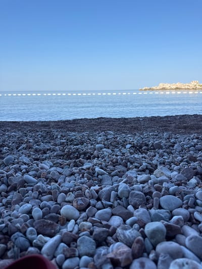 The image shows a close-up view of a pebble beach on a clear day in Sveti Stefan, Montenegro. The foreground is dominated by smooth, grey and brown pebbles of varying sizes. Beyond the pebbles, a dark strip of wet sand or smaller stones marks the high tide line. The tranquil sea, a calm blue, stretches to the horizon, where a white buoy line is visible. In the distance, a rocky island formation is partially submerged in the water. The sky is a clear, light blue, with no visible clouds. A sliver of a red object is visible in the bottom left corner of the frame.