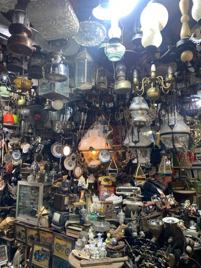 The image shows an interior scene of a cluttered shop in Surakarta, Indonesia, located at coordinates -7.569 latitude and 110.822 longitude. The shop is filled with an array of antique oil lamps, chandeliers, and various other trinkets, including cameras, framed photographs, and other miscellaneous objects. The ceiling is covered in a dense network of electrical wires and hanging fixtures. The shop is dimly lit and the atmosphere is somewhat chaotic.