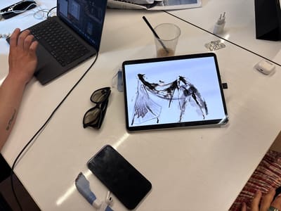An overhead, eye-level medium shot captures a white, slightly scratched tabletop. To the left, a person's left arm and hand are visible, their fingers resting on a laptop keyboard. The laptop is dark gray with a screen displaying code and design software. In the center of the frame, a tablet displays a black and white abstract digital painting. To the right of the tablet, a clear plastic cup with a straw contains a light brown liquid, likely iced coffee. Next to the cup is a blister pack of pills. Further to the right are white Apple AirPods in their charging case, and a white USB charger. Below the tablet are a pair of black-rimmed sunglasses and a black smartphone. To the bottom right, a person's hand, adorned with a green ring, is partially visible, resting on a striped fabric. The overall scene suggests a workspace. The prompt's request to specify the location in Milan, Italy, cannot be fulfilled as the image provides no geographical indicators.