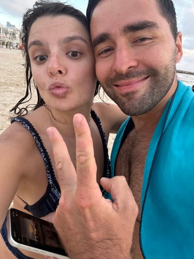 This media file is a selfie taken by two people on a beach, likely in Tel Aviv, Israel, given the geotag. The image is oriented vertically, with the subjects' faces filling the majority of the frame.
The person in the foreground on the right is wearing a dark bikini top with a patterned trim. Their hand is raised, and they are making a peace sign with two fingers. Their lips are pursed in a "duck face" pose. They have a nose ring and their hair is dark and appears wet.
The second person, positioned behind and to the left, is also looking towards the camera. They have dark hair and a beard, and are wearing a bright blue towel draped over their shoulders. Their eyes are open, and they have a slight smile.
The background shows a sandy beach with some faint structures and possibly people in the distance, suggesting an outdoor, daytime setting. The sky is overcast, indicating possibly cloudy weather. The time stamp on the phone visible in the lower right corner reads "19:21," suggesting it is late afternoon or early evening. The phone screen displays the word "Latest," possibly referring to a news feed or social media content. The overall mood appears casual and relaxed, consistent with a beach outing.