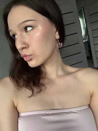 The image shows a close-up, rotated view of a young person's face and upper torso. The person has dark brown hair, which is partially obscuring their face. They are wearing a light pink strapless top and a small, heart-shaped pink earring in their left ear. They also have a nose ring. The background appears to be a textured surface, possibly a wall with vertical lines, and there's a metallic object with a light source visible in the upper portion of the image. The lighting suggests an indoor setting, possibly with artificial light. The person's skin appears smooth and well-lit, with some subtle highlights. The overall mood is casual and intimate due to the close-up perspective. There is no visible text in the image.