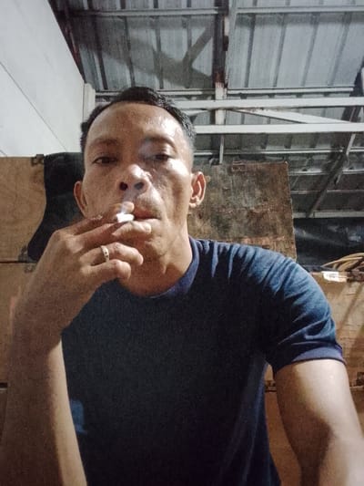 A close-up, over-the-shoulder selfie shows a middle-aged Indonesian man smoking a cigarette indoors. He is wearing a dark blue t-shirt and a ring on his left hand. The background appears to be a dimly lit storage area with wooden crates and exposed metal roofing. The man's face is turned towards the camera, and he is exhaling smoke. The image does not provide information about the specific location within Bukit Ketok, Indonesia.
