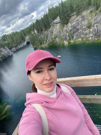 In the city of Ruskeala, Russia, a young woman with brown hair tied in a ponytail is taking a selfie on a wooden bridge overlooking a picturesque landscape. She is wearing a pink baseball cap, a white turtleneck, and a pink hooded sweatshirt. A beige backpack strap is visible over her shoulder.
The background reveals a stunning view of the Ruskeala Mountain Park, known for its marble canyon. A dark, serene lake fills the canyon, reflecting the surrounding lush green pine trees and the dramatic, rocky cliffs. The cliffs are characterized by their grey, textured surface, with some sections showing exposed white marble.
In the distance, a small boat with two people can be seen on the lake. A tunnel entrance is visible carved into the cliff face on the left side of the frame. Elevated wooden walkways with railings are constructed along the edges of the canyon, allowing visitors to traverse the area. The sky above is partly cloudy, with a mix of grey and white clouds against a pale blue backdrop. The overall scene captures the natural beauty and recreational aspects of the Ruskeala Mountain Park.