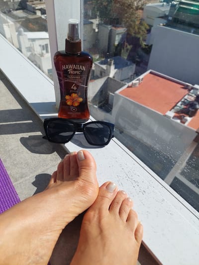The image captures a top-down view of a person's bare feet, a bottle of tanning oil, and sunglasses, all resting on a white, textured window ledge or balcony railing. The scene suggests relaxation and sunbathing on a bright day.
Two bare feet, with toes sporting a light, shimmery nail polish, are positioned towards the right of the frame. To their left, a dark brown bottle of Hawaiian Tropic tanning oil with a spray top lies horizontally. The label on the bottle is clearly legible, displaying "HAWAIIAN TROPIC™ TANNING ACEITE BRONCEADOR CON FILTRO SOLAR EN SPRAY," along with "4 SPF/FPS UVB," "BRONCEADO PERFECTO," "FORMULA HUMECTANTE," and "240 mL." Adjacent to the tanning oil is a pair of black sunglasses. A strip of a purple, textured mat is visible at the very bottom edge of the image.
Below the ledge, a cityscape is visible through a transparent surface, possibly glass or a clear barrier, which appears somewhat streaked or dirty. The background shows various buildings and rooftops, including one prominent structure with a reddish-orange roof, indicative of an urban setting. Bright, direct sunlight bathes the scene, casting sharp shadows from the feet, bottle, and sunglasses, confirming a sunny day, likely mid-morning or afternoon. The overall impression is one of leisure in warm weather, set within an urban environment in Buenos Aires, Argentina.