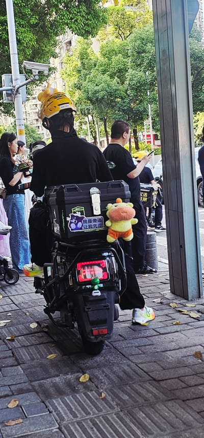 A medium outdoor shot, rotated counterclockwise 90 degrees, shows a group of people on the sidewalk of a street in the Yuzhong District of Chongqing, China. At the right is a brick sidewalk with scattered leaves. Moving left, a scooter is parked on the sidewalk, with two people standing near it. The scooter has a black box on the back with stickers and a stuffed animal. A third person is sitting on the box, looking at a phone, and a fourth person is standing next to the scooter, also looking at a phone. A yellow hardhat with a stuffed animal on top is visible. In the background are trees, buildings, and a gray metal structure overhead.