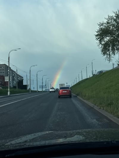 The image captures a segment of a road in Izhevsk, Russia, likely during the daytime, as indicated by the presence of a rainbow. The sky is overcast, suggesting recent or ongoing rain, which has left the asphalt road wet and reflective. A red car is visible on the road, traveling in the same direction as the viewer. Several streetlights line the side of the road, their curved posts creating a repeating pattern. Beyond the road, a grassy embankment slopes upwards, and further in the distance, apartment buildings are visible. A tree with dark foliage occupies the upper left corner of the frame. A faint rainbow arches across the sky, its colors muted by the cloudy conditions. There is no visible text in the image.