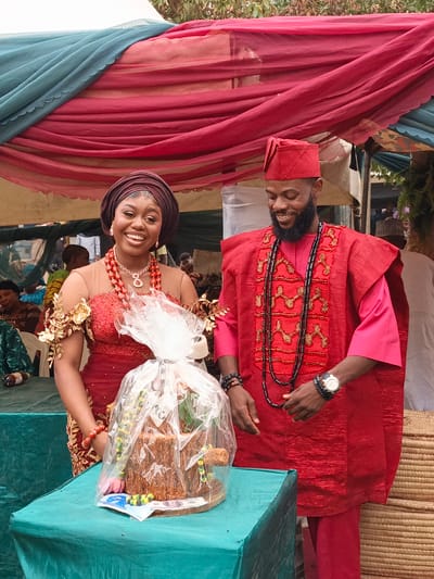 A joyous, daytime outdoor ceremony in Sabon Gari West, Nigeria, features a smiling man and woman in vibrant traditional attire. The woman wears a reddish-brown headwrap, a red and gold-embroidered dress with sheer elements, and elaborate red beaded jewelry, including a prominent necklace and earrings. The man is dressed in a red tunic (agbada-like), trousers, and cap, adorned with gold and black embroidery and multiple long black beaded necklaces. He also wears a silver watch and beaded bracelets.

The couple stands in front of a teal-covered table where a gift, possibly a cake or symbolic item, is presented. The gift is wrapped in clear plastic with a silver ribbon, and Nigerian Naira banknotes (e.g., ₦200, ₦1000) are placed near it, with visible text "CENTRAL BANK OF NIGERIA". The background shows an outdoor setting, likely under a canopy, decorated with red, teal, and beige draped fabrics. Other attendees are partially visible in the softly focused background. Both individuals display clear emotions of happiness and celebration, with the woman's wide smile and the man's gentle grin. The bright natural lighting indicates it is daytime. The overall scene conveys a festive and culturally rich event.
