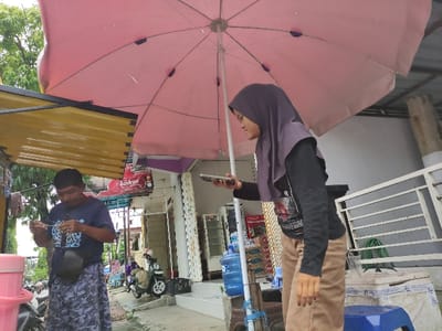 This media file captures an outdoor scene, likely a street or market area, in Situbondo, Indonesia, during the daytime. The foreground is dominated by a large, open pink market umbrella, suggesting sunny weather.

Two individuals are visible. On the left, a woman wearing a dark long-sleeved top, light brown pants, and a purple hijab is holding a smartphone in her left hand, appearing to be taking a photo or video, or checking the screen. Her right arm is raised. On the right, a man in a dark blue t-shirt with a white design and camouflage-patterned shorts is holding a small object or device in his hands, looking down at it. He also wears a dark bag slung across his shoulder.

In the background, various objects and structures are visible, including a blue water dispenser bottle on a stand, shelves stocked with packaged goods (possibly snacks), and a small white metal rack. Further back, a child's ride-on toy vehicle is parked near several buildings that appear to be residential or small shopfronts. A yellow corrugated metal structure, possibly part of a stall roof, is seen on the right. Green foliage is visible in the upper right corner.

Notable visible text includes "REACH" on the man's blue t-shirt and "Bodrex" (a brand name) on a red banner or sign in the mid-ground. The scene depicts a casual, everyday street activity in an Indonesian setting.