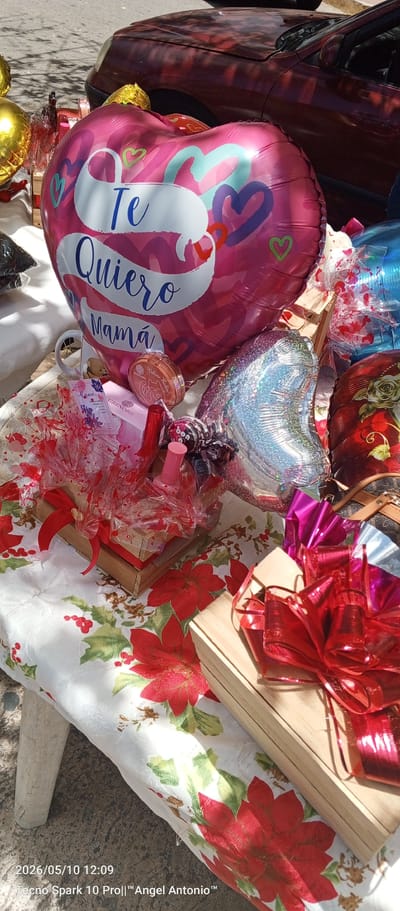 The image shows a display of gifts, presumably for Mother's Day, set up on a table covered with a white tablecloth with red poinsettia designs. The gifts include multiple heart-shaped balloons, one pink with the Spanish words "Te Quiero Mamá" (I Love You Mom), another silver, and others with various patterns. There are also wooden boxes adorned with red bows, filled with various items such as makeup and nail polish. A white mug with a teddy bear design is visible behind the pink balloon. A small handbag with a patterned design is also present.
In the background, there is a maroon car parked on what appears to be a street. The image was captured on May 10, 2026, at 12:09, using a Tecno Spark 10 Pro phone. The location, Juan Griego, Venezuela, is not evident from the image itself but is provided as external information.