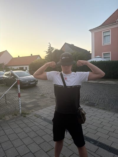 A young person, wearing a white t-shirt with black Adidas stripes, black shorts, and a dark baseball cap, flexes their bicep. The person is standing on a paved sidewalk in front of a residential building. The t-shirt has a prominent Adidas logo on the chest and repeating Adidas text along the sleeve. The baseball cap has the Alpha Industries logo visible. A small, brown pouch is worn crossbody. The setting appears to be a suburban street with houses and a car visible in the background. The pavement is made of grey cobblestones. A metal fence with red and white markings is in the foreground. The image is taken outdoors during what appears to be late afternoon or early evening, with a warm, soft light illuminating the sky. The weather seems clear and calm. The license plate of the car is visible and reads "NI-CO 201".