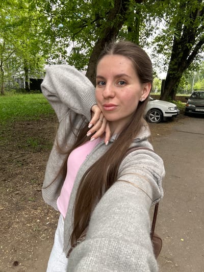 A young woman with long brown hair and light skin is posing for a selfie outdoors. She is wearing a gray zip-up hoodie over a pink top, and white pants. Her head is resting on her hands, with a ring visible on her left ring finger.
The background includes green foliage, suggesting a park or garden setting. A road and parked cars are visible in the distance. Based on visual clues, the location is Klin, Russia. The weather appears overcast or partly cloudy.