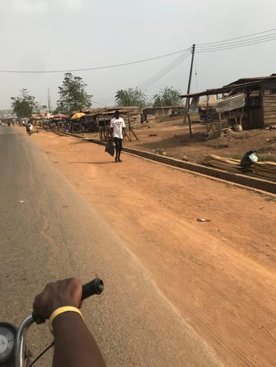 The scene depicts a street view in Ogbese, Nigeria, under daylight conditions. The foreground features the right hand and forearm of an individual operating a motorcycle or similar vehicle, gripping a black handlebar. A yellow wristband is present on the subject's left wrist. The immediate ground consists of an asphalt road surface transitioning to a reddish-brown dirt shoulder on the right.

In the midground, a dark-skinned male subject walks from left to right on the dirt shoulder. He wears a white t-shirt with a multi-colored graphic on the chest (four playing card suits), dark shorts, and dark footwear. He carries a black bag in his left hand. A distinct shadow extends from his left side onto the dirt. The dirt shoulder displays tire tracks and scattered debris. A concrete curb separates the paved road from the dirt shoulder in the midground.

The background reveals a series of rudimentary roadside structures, constructed primarily from wood and corrugated metal, consistent with market stalls or small shops. Some structures feature fabric awnings. A tall wooden utility pole with multiple overhead wires is visible on the right side. Piles of rough-cut wooden planks are stacked near one of the structures on the far right. Further down the road on the left, additional vehicles and indistinct figures are present. The sky above is light and hazy. No direct interactions between individuals are discernible.