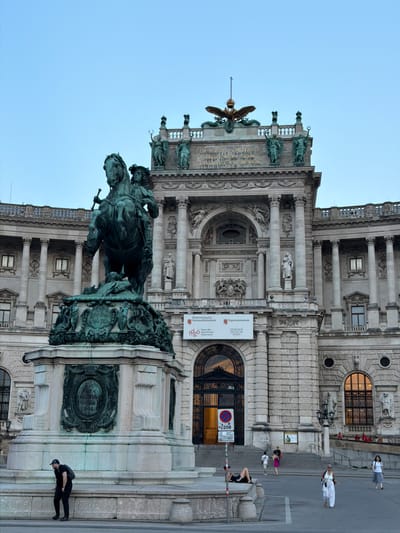 The image depicts the equestrian statue of Archduke Charles in front of the Neue Burg wing of the Hofburg Palace in Vienna, Austria. The statue, cast in bronze, is mounted on a large stone pedestal adorned with relief sculptures. A man wearing a black cap, black t-shirt, and black pants with a backpack is bending down near the base of the monument. In the background, the grand facade of the Neue Burg, a historical imperial palace, is visible with its classical architecture, columns, and statues. Several people are seen walking on the pavement and stairs in front of the palace, and some are seated on the steps. Banners advertising museums are displayed on the building's entrance. The sky is clear and light blue, suggesting it is either early morning or late evening.