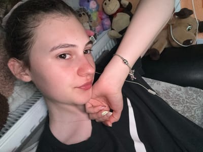 In a domestic indoor environment, a young person with dark hair pulled back in a bun lies down, resting their head on their arm. They are looking directly at the camera with their eyes open. Their left ear is visible, and they are wearing a thin silver chain necklace. Their right arm is bent, with their hand resting near their face. A beaded bracelet with charms adorns their wrist. Their fingernails are painted with a light yellow polish and some have green polka dots. They are wearing a black t-shirt. In the background, several stuffed animals are visible, including a brown cow, a monkey, and other colorful toys. The scene appears to be set in Panagyurishte, Bulgaria, within a bedroom or living area, suggested by the presence of a radiator under a window.