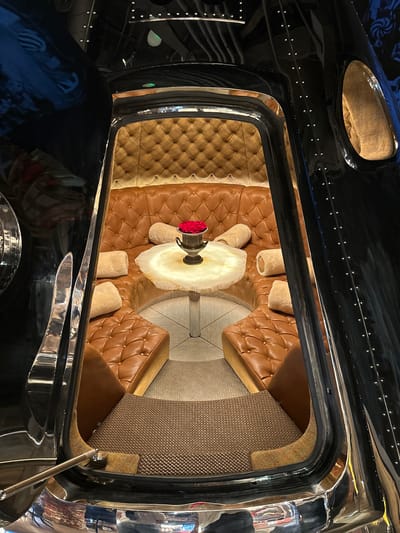 The image displays the interior of a luxurious, custom-designed seating area, possibly within a vehicle or a themed establishment. The space is characterized by deep, button-tufted brown leather upholstery, forming a curved couch and individual seats. A small, translucent table with a rough, natural edge sits in the center, adorned with a silver vase holding a bouquet of red roses. Round, fluffy pillows are scattered across the seating. The exterior of this enclosed space is glossy black, with visible metallic rivets and curved metallic trim, suggesting a high-end, bespoke construction. The flooring is a textured, dark grey carpet. The overall aesthetic evokes a sense of opulent privacy and retro-futurism. The location is identified as London, United Kingdom. There are no people visible in the image.