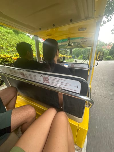 In the city of Tây Hoa Lư Ward, Vietnam, a yellow open-air electric vehicle, likely a tourist buggy, is captured in motion on a paved road. The vehicle's interior is visible, revealing a driver and a passenger seated in front, both facing forward. The driver, a person with dark hair, is operating the vehicle, while the passenger, also with dark hair, is looking ahead. In the foreground, the legs of two individuals are visible, suggesting they are passengers in the rear of the buggy. One person wears dark green shorts, and the other wears a green garment. The buggy has a white panel affixed to its rear, displaying QR codes and text in both Vietnamese and English, indicating a feedback mechanism for the "Bai Dinh Spiritual and Cultural Tourism Complex." The surrounding environment features lush green foliage, including trees and bushes, indicating a natural or park-like setting. The sky appears overcast. The overall scene conveys movement and a tourist experience within a scenic, likely cultural or spiritual, destination.