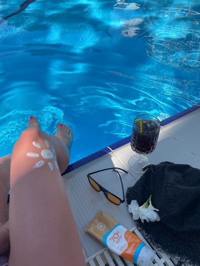 This media file captures a relaxing poolside scene in Alanya, Turkey. The main subject is a person's legs and feet as they dip into a bright blue swimming pool. Sunscreen, shaped into a sunburst, is visible on one leg, and bright pink nail polish adorns the toes.
Surrounding the pool edge, several items suggest a leisurely day: a pair of sunglasses, a dark bucket hat with a white flower placed on it, and a glass containing a dark beverage with ice and a yellow stirrer. A tube of sunscreen, labeled "70+ SPF ULTRA PROTECTION," is also positioned nearby.
The water in the pool reflects sunlight, creating shimmering patterns. The poolside tiling is a light, textured material. The overall impression is one of a warm, sunny day, ideal for swimming and relaxing. The presence of sunscreen and a cooling drink suggests a typical vacation or leisure activity. The time of day appears to be daytime, with the bright sunlight illuminating the scene.
