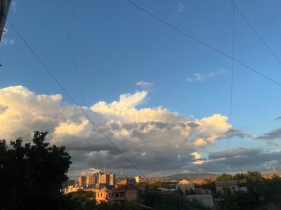 The image captures a view of Yerevan, Armenia, during what appears to be late afternoon or early evening. The sky is a dynamic mix of bright blue and scattered clouds, some of which are illuminated by the setting sun, creating a warm, golden glow against darker, more shadowed clouds. Overhead, a network of dark utility wires crisscrosses the frame, a common feature in urban landscapes.

In the foreground, on the left, the silhouette of a large, dense tree partially obscures the view. Below and extending to the right, a cityscape unfolds. Various residential buildings, characterized by their terracotta roofs and light-colored facades, are visible. Further in the distance, taller apartment buildings, typical of Soviet-era architecture, dominate the skyline, their facades catching the warm light. Beyond the urban sprawl, rolling hills and mountains form the backdrop, suggesting a natural landscape surrounding the city. The overall scene conveys a sense of a typical urban environment in Yerevan, with a mix of residential architecture, infrastructure, and natural topography. There are no visible people or animals in the image, and no specific actions or interactions are depicted.