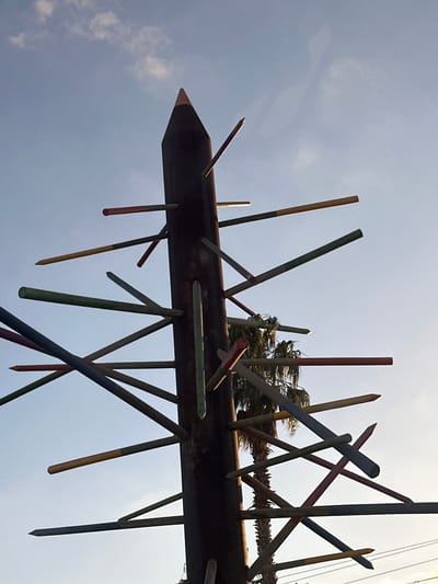 The image shows an outdoor art installation resembling a giant pencil with numerous colored dowels or sticks protruding from its sides at various angles. The installation is positioned against a bright, slightly cloudy sky. The central object is a tall, dark, cylindrical structure with a conical top, mimicking the shape of a pencil. The colored sticks, which appear to be made of wood and painted in hues of red, yellow, green, blue, and white, are inserted into holes or slots along the main structure. Some of the sticks have sharpened points, resembling pencil tips. In the background, the fronds of a palm tree are visible, suggesting a warm climate. The overall impression is one of a whimsical, large-scale sculpture. This art installation is located in the city of Petah Tikva, Israel.