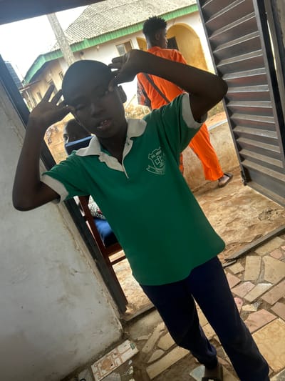 The image shows a young boy in a green polo shirt and navy blue pants standing in an outdoor setting. He is making a playful gesture, with his fingers near his eyes. Behind him, another boy wearing an orange outfit is visible, and further in the background, there are buildings with green roofs and arched doorways, indicative of a Nigerian setting. The ground is paved with broken tiles and dirt. The overall scene suggests a casual, everyday moment in a neighborhood within Ikorodu, Nigeria.