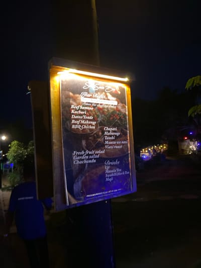 The image shows a menu board illuminated by a strip of light above it. It is mounted on a wooden post and is positioned on a sidewalk at night. The menu is for a restaurant called "Spice Bay." It advertises a "flair buffet menu" of various dishes, including beef samosa, kachori, dates/tende, beef makange, BBQ chicken, chapati, maharage, tambi, meusu wa nazi, viazi roast, fresh fruit salad, garden salad, chachandu, uji, masala tea, Swahili black tea, and maji. The image also shows a person in a blue shirt walking away from the menu, passing on the left side. This scene is located in Dar es Salaam, Tanzania, at the coordinates -6.754, 39.248.
