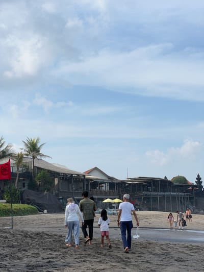 A diverse group of people are walking on a sandy beach in front of a row of restaurants and bars with palm trees in the foreground. The sky is partly cloudy, with a gentle breeze implied by the movement of the clouds. The beach is mostly wet and sandy, with some debris and footprints visible. The buildings are modern with open-air seating and umbrellas. There is a red flag with a symbol of a swimmer and a circle with a diagonal line through it, indicating no swimming. The people are dressed in casual beachwear, and some are carrying bags or other items. The scene appears to be in North Kuta, Indonesia, a popular tourist destination known for its beaches and nightlife.