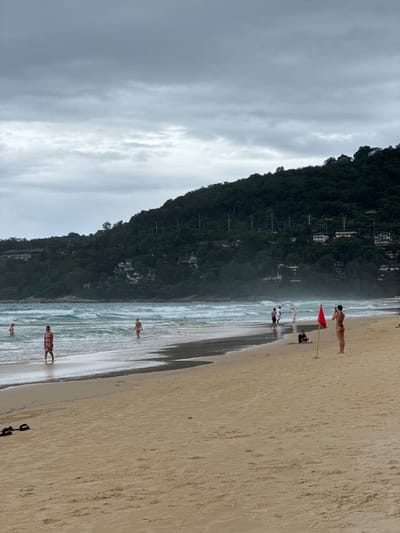 The image depicts a beach scene in Karon, Thailand. The foreground is dominated by a sandy beach, with gentle waves lapping at the shore. A lush, green hillside covered in trees rises steeply from the back of the beach, with some buildings visible nestled amongst the foliage. The sky is overcast, suggesting a cloudy or possibly rainy day.

Several people are present on the beach and in the shallow water. In the middle ground, a woman in a bikini stands on the sand, appearing to take a photo with her phone. Nearby, a red flag on a yellow pole is planted in the sand, possibly indicating a swimming area or a warning. Other individuals are scattered along the shoreline, some wading in the water and others walking on the sand. One person is visible near the water's edge, appearing to be wearing red and white patterned swimwear. Further down the beach, another individual is seen wading in the ocean.

The overall atmosphere appears relaxed, with people enjoying a day at the beach. The cloudy sky might suggest cooler temperatures or a break in sunny weather, but the presence of people in swimwear indicates it's still warm enough for beach activities. The dense vegetation on the hillside is a characteristic feature of tropical coastal areas. No visible text is present in the image.