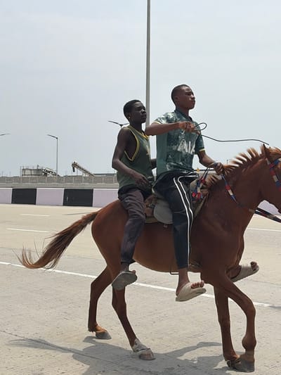 Two individuals are riding a single bay horse on a paved road surface in Igboefon, Nigeria, under a bright, clear sky. The front rider, a male, wears a dark green t-shirt with a white pattern, black track pants with white side stripes, and light-colored sandals. He is positioned on a light-colored saddle, holding dark reins in his right hand. The second rider, a male, is seated behind the first, wearing a green tank top with yellow trim, dark pants, and grey sandals. Both individuals are looking forward and slightly to their right. The brown horse has a darker mane and tail and is equipped with a bridle featuring red, blue, and white decorative elements. The horse's legs are in mid-stride, indicating movement. The road surface is grey with a visible white line. In the background, a black and white striped concrete barrier is present. Beyond the barrier, industrial structures including a conveyor system and concrete installations are visible under the bright sky, along with two tall street light poles.