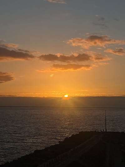 The image depicts a sunset over the ocean. The sky is a mix of orange, gray, and light blue with clouds. The sun is visible as a bright orange orb on the horizon. The ocean is reflecting the sunlight, creating a sparkling effect. A dark, rocky shoreline is visible on the right side of the image. The image was taken in Mogán, Spain.