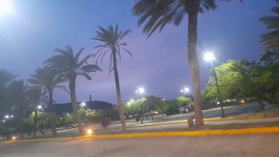 A wide shot captures a twilight scene in Pedro González, Venezuela, with a clear sky transitioning from deep blue to a soft purple. The foreground is a light brown, paved surface, likely a road or parking lot, bordered by a bright yellow line. Beyond this, a strip of green grass separates it from a road where several vehicles, including a white SUV, a black SUV, and a dark-colored car, are parked or in motion. Tall palm trees dominate the midground, their fronds silhouetted against the sky. Some of these palm trees are illuminated by streetlights that cast bright white beams. In the background, a cluster of green trees and some lower vegetation are visible. Further in the distance, on a gentle hill, a lighthouse with black and white horizontal stripes stands prominently. The overall atmosphere is serene and peaceful, with the artificial lights creating a warm glow amidst the natural dusk.