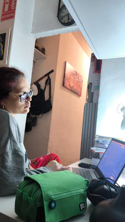 The scene depicts an interior setting, identified as a location within the city of Ripollet, Spain. A woman with dark hair and glasses is seated in profile, facing right, looking intently at an open silver laptop positioned on a light-colored desk. She is wearing a grey long-sleeved top and red patterned pants; her right hand is situated near the laptop's keyboard. In the immediate foreground, a bright green canvas bag with a flap closure, a blue and white circular zipper pull, and a white "Big Dudes" tag is visible on the desk surface. To the right of the bag are a black computer mouse and black wired headphones. A black smartphone rests on the desk next to the laptop. A stack of papers is present beside the laptop, and a white desk lamp with an illuminated bulb is visible to the far right. The background features light-colored walls. On the upper left wall, a red "EXTINTOR" (fire extinguisher) sign is mounted above a partially visible framed artwork. High above, a round, dark-framed wall clock displays a time around 5:30. An architectural opening leads to a further area where a wall-mounted picture, shelves containing various objects, a hanging black backpack, and a dark stick-like object are discernible. To the far right in the background, a vertical blind is visible.