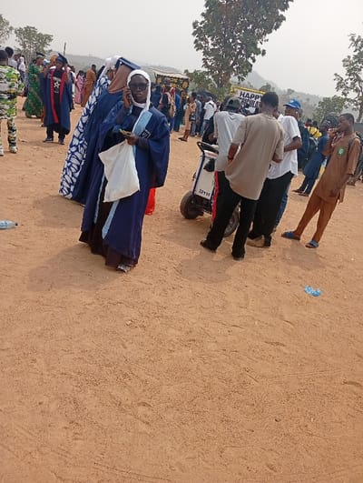 The image depicts an outdoor gathering in Adankolo, Nigeria, on a dry, reddish-brown dirt ground under an overcast or hazy sky. In the foreground-left, a female wears dark blue academic regalia with light blue trim, a white head covering (hijab), and dark sunglasses. She holds a white plastic bag in her left hand and a small yellow object, while her right hand holds a mobile phone to her ear. She wears patterned dark sandals. To her immediate left on the ground, a discarded plastic water bottle is visible. Further back, another individual is seen in similar dark blue academic regalia with a blue and white patterned fabric draped over their shoulder.
In the mid-ground, a group of males stands around a white and black handcart or trolley with two wheels, marked "3211/4." One male in a light brown short-sleeved shirt faces away from the camera. Another male in a white shirt and blue jeans stands beside him. To the right, a male in a light brown traditional outfit and blue sandals stands, looking towards the left foreground, also holding a mobile phone to his ear. A crumpled blue plastic wrapper lies on the dirt ground in the foreground-right.
The background shows a large crowd of people, some wearing academic gowns and caps, others in various casual and traditional attire. Partially visible banners in the background include text such as "HAPPY R...TION" and "WELCOME TO THE...". Sparse trees and distant hills are visible on the horizon. A person in camouflage-patterned clothing is visible on the far left. The scene suggests a public event or ceremony.