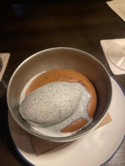 A close-up, overhead shot shows a serving of dessert presented in a silver bowl. The dessert consists of a round, brown cake or pastry topped with a large scoop of light grey ice cream speckled with black. A thin layer of white sauce or cream surrounds the base of the ice cream and also covers parts of the cake. The bowl sits on a white plate with a thin blue rim, which is partially visible. Underneath the dessert bowl, a small, folded piece of brown paper is tucked into the side. The background is dark and out of focus, with hints of a dark wooden table and a white placemat or napkin. The lighting suggests it might be indoors, possibly in a restaurant setting, and the time of day is unclear due to the lack of natural light cues. There are no people visible in the frame. The focus is entirely on the dessert.