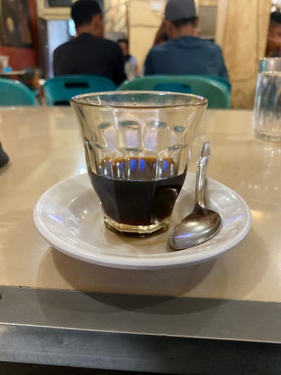 The image shows a close-up view of a glass of dark coffee on a white saucer, with a small silver spoon resting on the saucer. The coffee is dark brown and fills about a third of the glass. The glass has a textured surface and is transparent, allowing the reflection of blue and white light from above. The saucer is white and circular, with a slightly raised edge. The spoon is also silver and appears to be slightly tarnished.
The scene is set on a table with a light-colored, speckled surface, possibly a countertop or a table in a cafe. In the background, blurred figures of people are visible, seated at tables. The person closest in the background to the left is wearing a dark shirt. To the right, another person is wearing a dark blue shirt and a grey cap. There are also hints of green and turquoise chairs in the mid-ground, suggesting a public seating area. A glass of water is partially visible in the far right background.
The overall ambiance suggests a casual cafe or eatery, likely a local establishment. Considering the reference to Lhokseumawe, Indonesia, this scene could depict a coffee shop within the city, where locals gather to enjoy coffee. The setting appears to be indoors, with artificial lighting contributing to the reflections in the coffee glass. The image is focused on the coffee and its accompaniments, with the background providing context.