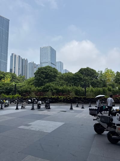 This image depicts an outdoor scene in Tianhe District, China, featuring a row of parked electric scooters and motorcycles in the foreground. In the middle ground, a dense line of lush green trees forms a natural backdrop, obscuring the lower portions of several tall, modern buildings that rise into the sky. Two individuals are walking in the mid-ground, one holding a white umbrella. The ground is paved with large, grey stone tiles. The sky is overcast with scattered clouds.