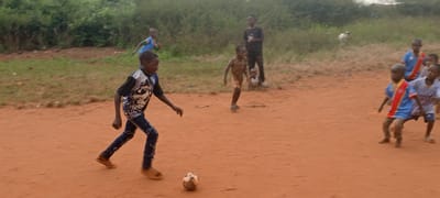 Children play soccer on a dirt field in Ngalula, Democratic Republic of the Congo. In the foreground, a boy in a skull-print shirt and torn jeans dribbles a soccer ball. Other children are scattered around the field; several wear soccer jerseys, while one stands in the background holding a ball. The background features green vegetation and scrubby ground.