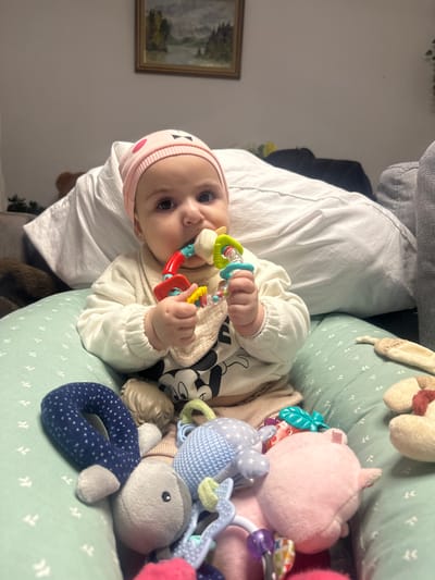 A baby wearing a pink hat and beige outfit lies on a green patterned surface, surrounded by toys. The baby holds a colorful teether in their hands, with one end in their mouth. Stuffed animals, including a pink pig and a blue whale, are scattered around the baby. A white pillow supports the baby. A framed landscape picture hangs on the wall in the background. The lighting is soft, and the overall scene appears cozy.