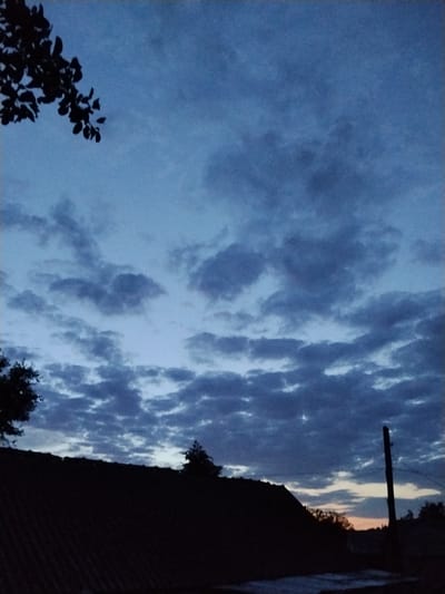 The image depicts a twilight sky over Ustyvytsia, Ukraine. The sky is a deep blue, transitioning to a lighter hue near the horizon, with scattered clouds. In the foreground, a dark silhouette of a roofline is visible, with a tree branch extending from the upper left corner. A dark, vertical pole stands on the right side of the frame. The overall scene is one of dusk or dawn in a semi-urban or rural setting.