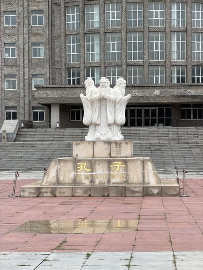 In front of a large, imposing building with a grey brick facade and numerous windows, a monument stands on a stepped stone platform. The monument itself is a white sculpture of three figures, appearing to be ancient scholars, with long beards and flowing robes. They are positioned in a circular arrangement, facing outwards. The platform beneath the sculpture has inscriptions in gold Chinese characters. The ground in front of the monument is paved with reddish-brown tiles, with some puddles of water reflecting the surroundings. A wide set of stone stairs leads up to the building's entrance, which features glass doors. The scene appears to be an outdoor public space, possibly a university campus or a cultural landmark. The building is identified by Chinese characters on its facade as the "Chonglou" (存楼), which translates to "Storage Building" or "Archives Building". This building is located at the Shenyang Institute of Engineering in Shenyang City, China.