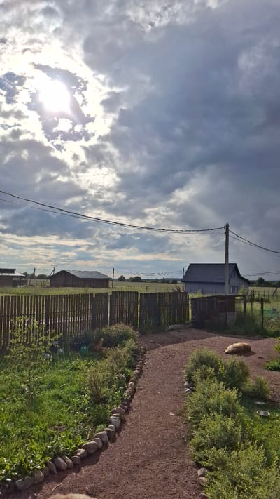 The image depicts a rural scene in Kempelevo, Russia, under a dramatic sky with bright sun peeking through dark clouds. A gravel path leads into a yard bordered by green grass and bushes. A wooden fence runs across the middle ground, separating the yard from a grassy field containing a dark wooden structure and a light-colored house with a dark roof. Overhead, power lines crisscross the sky. In the yard, a light-colored dog is lying down near a bush.